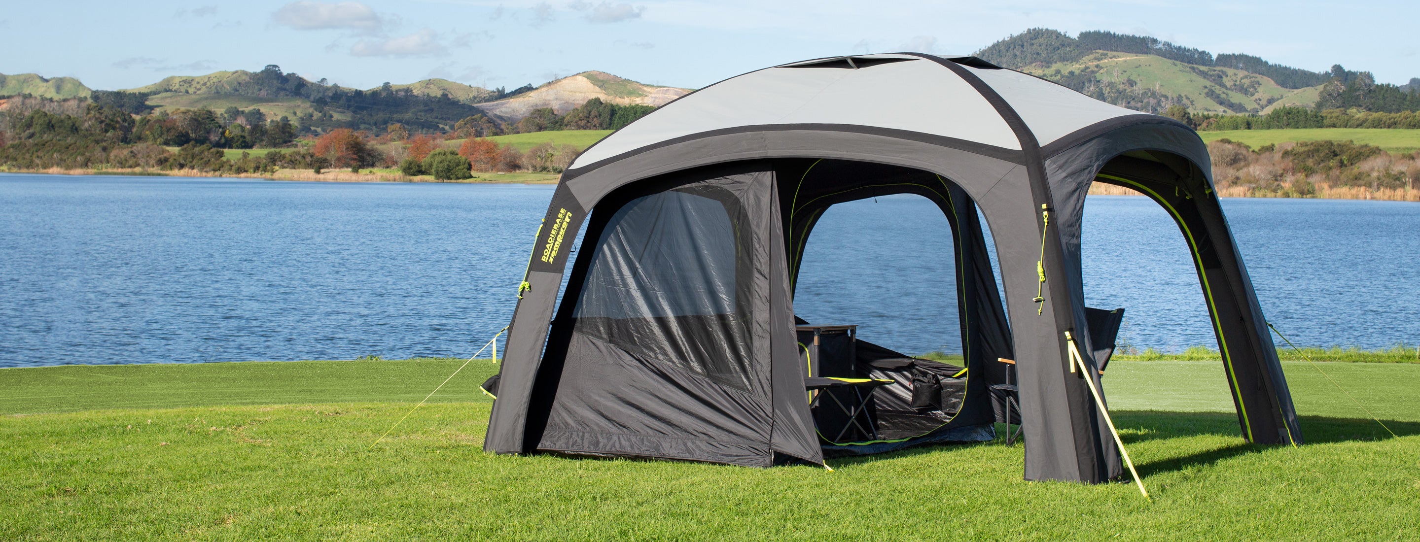Outdoor canopy tent set up on grass near a body of water with scenic hills in the background.
